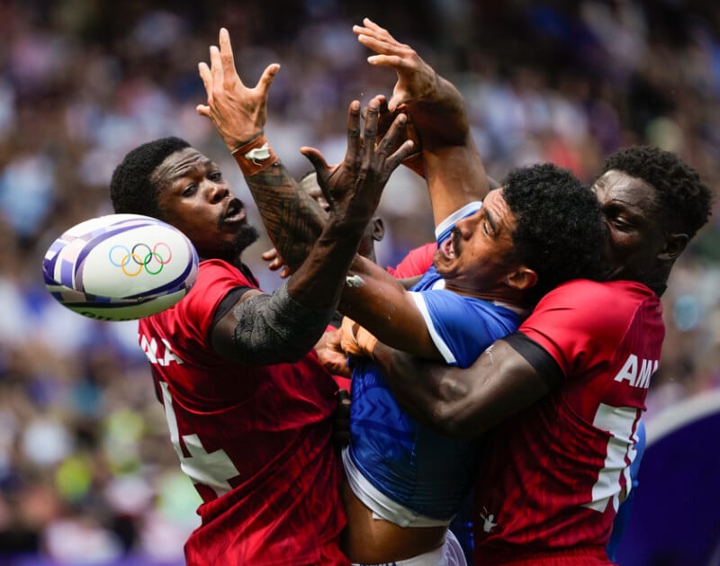 Rugby players from two teams fiercely compete for the ball mid-air. Three players in red jerseys and one in a blue jersey are intensely focused, with arms outstretched, as they try to gain possession. The scene captures the action and energy of the match.