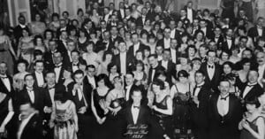 A black and white photo of a large group of people at a formal gathering in 1927. Many are in tuxedos and evening dresses, smiling and facing the camera. The setting suggests a grand ballroom atmosphere.