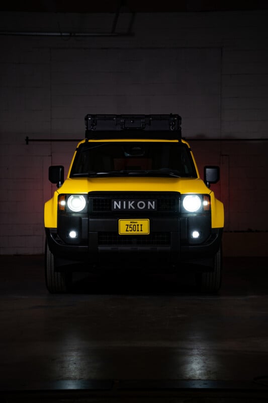 A bright yellow SUV with "NIKON" on its front, headlights on, parked indoors against a white brick wall. A yellow license plate reads "2501," and a roof rack is mounted on top.