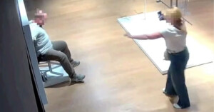 A security camera image shows a person sitting on a chair while another person stands nearby pointing a phone at them. Both faces are blurred, and the scene takes place in a well-lit indoor space with wooden floors.
